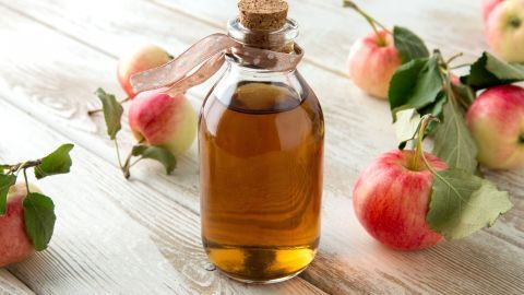 Eine Flasche Apfelessig steht auf einem Holztisch, umgeben von frischen, roten Äpfeln und grünen Blättern.