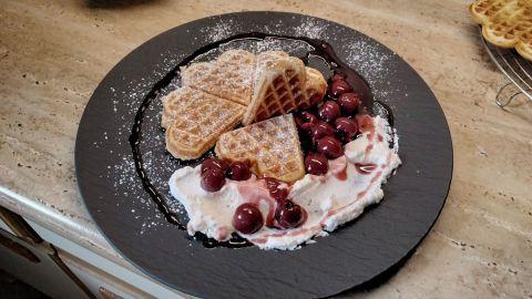 Waffeln liegen auf einem schwarzen Teller, garniert mit Kirschen und Sahne, umgeben von einem rustikalen Umfeld.