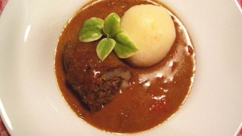 Ein weißer Teller enthält eine Portion Bratensauce, ein gefülltes Rinderstück und einen Kartoffelknödel, garniert mit einem Blatt.