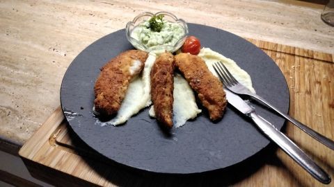 Panierte Hähncheninnenfilets liegen auf cremigem Kartoffelpüree, serviert mit einem frischen Salat und Tomate.