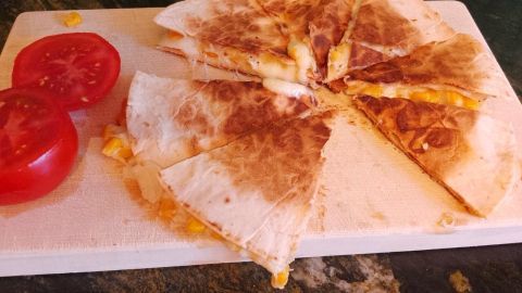 Käse-Quesadillas auf einem Brett, geschnitten in Stücke, daneben frische Tomatenscheiben, alles auf einer dunklen Fläche.