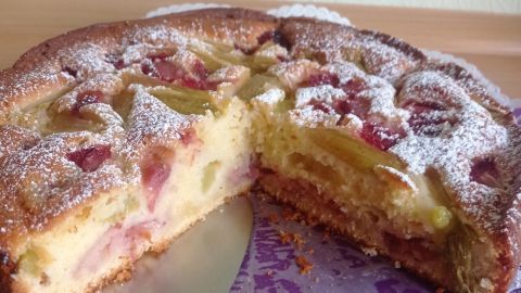 Ein Erdbeer-Rhabarber-Kuchen mit Puderzucker bestäubt steht auf einer lila serviette, ein Stück ist mit einem Tortenheber herausgeschnitten.