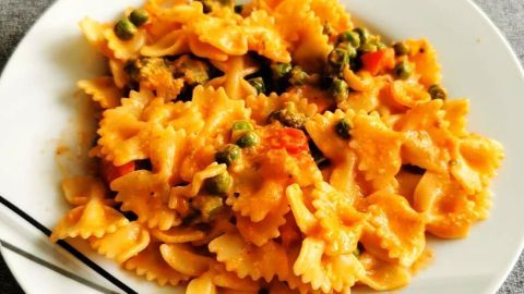 Farfalle in einer cremigen Tomaten-Sahnesoße liegen auf einem weißen Teller, umgeben von einem grauen Untergrund.