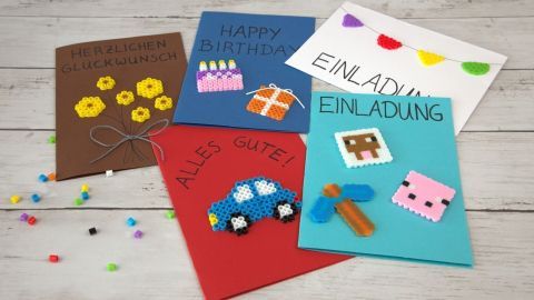 Bunte Karten mit Bügelperlen zeigen Grüße und Einladungen, kreativ gestaltet auf einem hellen Holztisch.