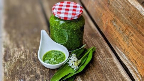 Ein Glas Bärlauchpesto steht auf rustikalem Holz neben einer kleinen Schale mit Pesto und frischen Bärlauchblättern.