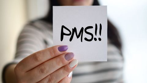 Eine Hand hält einen Zettel mit der Aufschrift „PMS!!“ vor einem verschwommenen Hintergrund, was die Thematik des PMS verdeutlicht.