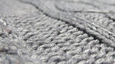 Grauer Strickstoff mit sichtbaren Fusseln und Knoten, der eine unebene Struktur zeigt und zum Kleidungsstück gehört.
