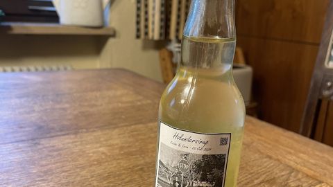 Eine Flasche Holunderblütensirup steht auf einem Holztisch, daneben ein Holzfahrrad, umgeben von einer gemütlichen Wohnatmosphäre.