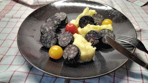 Auf einem Teller liegt cremiges Kartoffelpüree neben Scheiben von gebratener Speckwurst, umgeben von bunten Tomaten.