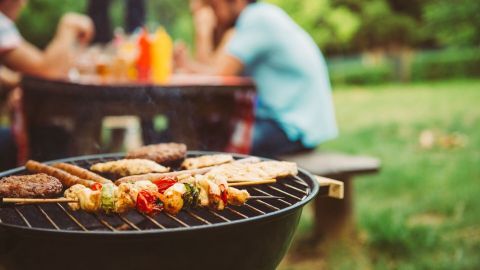 Auf dem Grill liegen verschiedene Fleischsorten und Gemüse, während im Hintergrund eine gesellige Grillrunde im Freien stattfindet.