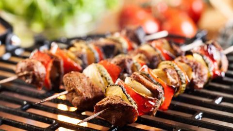 Gemüse-Fleisch-Spieße braten auf dem Grill, umgeben von frischem Gemüse und rohen Tomaten im Hintergrund.