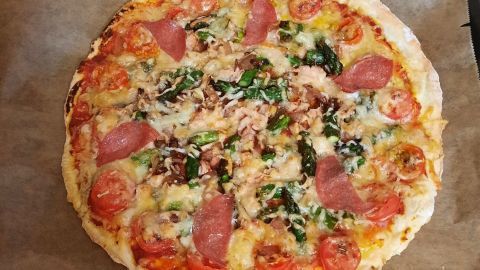 Eine frisch gebackene Pizza mit grünem Spargel, Tomaten und Schinken liegt auf Backpapier in einem Ofen.