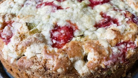 Ein frischer Rhabarber-Erdbeer-Kuchen mit knusprigen Streuseln liegt in einer runden Form auf einem Holzuntergrund.