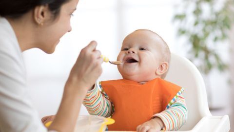 Eine mütterliche Figur füttert ein fröhliches Baby in einem Hochstuhl mit einem Löffel, während Licht durch ein Fenster strömt.