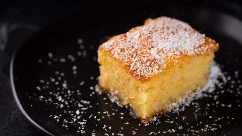 Ein Grießkuchen liegt auf einem schwarzen Teller, mit Kokosraspeln bestreut, in einer dunklen Umgebung.