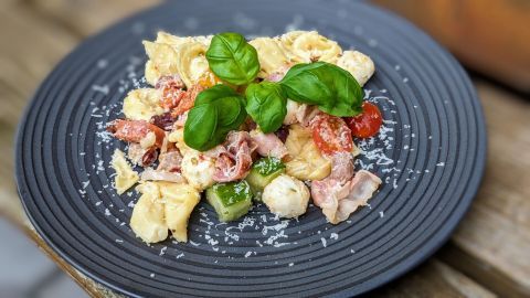 Auf einem tiefen, schwarzen Teller liegt ein bunter Tortellini-Salat mit Bacon, Mozzarella, Tomaten und frischem Basilikum.