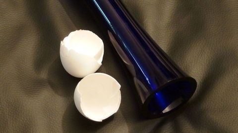 Schmutzige Vasen kann man sehr gut mit lauwarmem Wasser und Eierschalen reinigen. Eine blaue Vase steht neben zwei zerbrochenen Eierschalen auf einem weichen, dunkelgrünen Untergrund.
