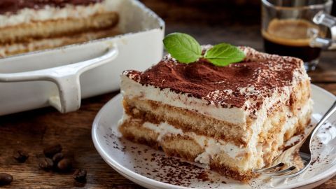 Ein köstliches Stück Tiramisu liegt auf einem Teller, umgeben von Kaffeebohnen und einer Tasse Kaffee.
