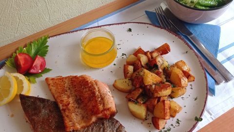 Lachsforellen-Filet liegt gebraten auf einem Teller, umgeben von Kartoffeln und frischem Salat, mit Zitronenscheiben.