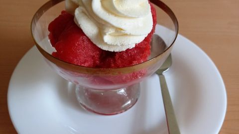 Ein Glas mit Erdbeer-Sorbet wird mit Sahnehaube und einer frischen Erdbeere dekoriert. Der Untergrund ist Holz.
