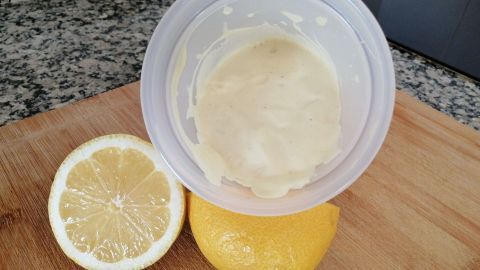 Eine weiße Knoblauchmayonnaise (Aioli) ohne Ei befindet sich in einer Schüssel auf einem Holzbrett, daneben liegen zwei Zitronenhälften.