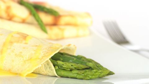 Ein gefüllter Crêpe umhüllt frischen Spargel und liegt auf einem weißen Teller, im Hintergrund weitere Crêpes und ein Besteck.