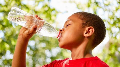 Ein Junge trinkt aus einer Wasserflasche, umgeben von einem grünen, sonnigen Baumhintergrund.
