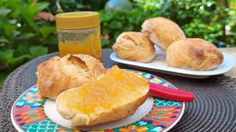 Ein knuspriges Brötchen mit Ananas-Zitronen-Gelee liegt auf einem bunten Teller, während weitere Brötchen im Hintergrund stehen.