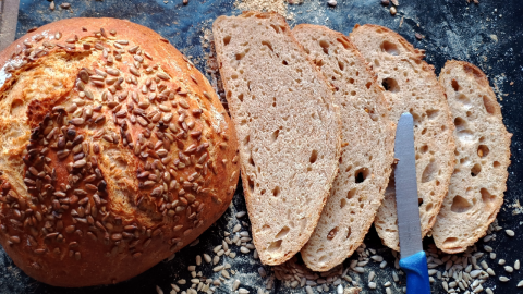 Ein runder, mit Sonnenblumenkernen bedeckter Joghurt-Brotlaib liegt neben mehreren geschnittenen Brotscheiben auf einer Arbeitsfläche.