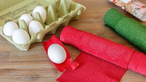 Weiße Ostereier liegen neben Rot- und Grüntönen von Krepppapier auf einem Holzuntergrund, bereit zum Buntfärben.