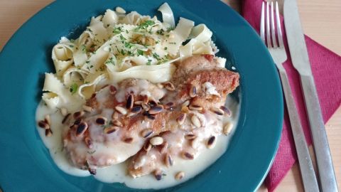 Eine zarte Hähnchenbrust mit Zitronen-Thymian-Sauce liegt auf einem blauen Teller, umgeben von bandnudeln und einer Schüssel Suppe.