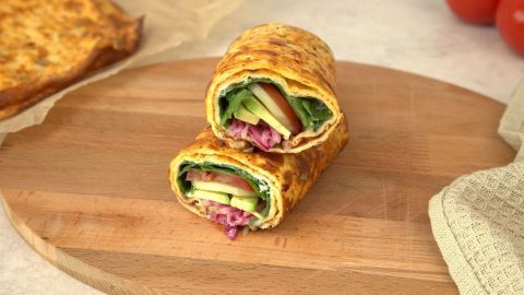 Auf einem Holzbrett liegen zwei gefüllte Hüttenkäse-Wraps mit Gemüse, umgeben von einer entspannten Küchenumgebung.