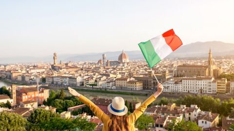 Eine Frau mit einem Hut hält eine italienische Flagge hoch und blickt auf die Stadt Florenz bei Sonnenuntergang.