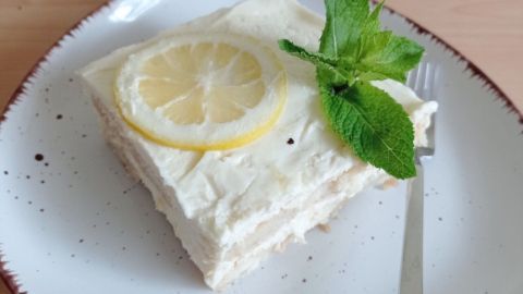 Eine cremige Zitronen-Tiramisu-Quadrat auf einem Teller, dekoriert mit einer Zitronenscheibe und frischer Minze, liegt auf einem Holztisch.