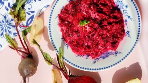 Ein bunter Rote Beete-Salat liegt auf einem Teller, umgeben von frischen Rüben und Apfelscheiben auf einem hellen Hintergrund.