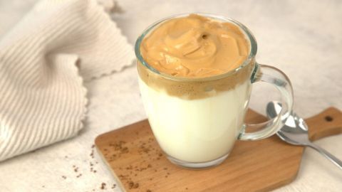 Ein Glas Dalgona Kaffee steht auf einem Holzbrett, oben schaumige Kaffeeschicht, daneben ein Löffel.