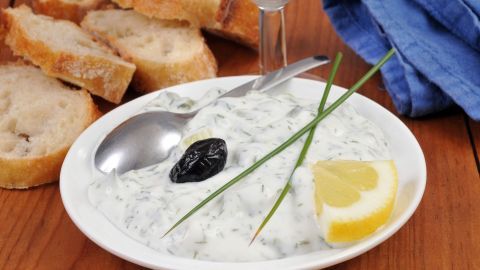 Eine Schüssel Tzatziki mit Kräutern und einem Spritzer Zitrone wird auf einem Holztisch serviert, umgeben von frischem Brot.