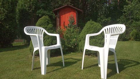 Zwei weiße Kunststoffgartenstühle stehen auf dem Rasen, umgeben von Sträuchern und vor einem roten Gartenhaus.