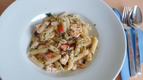 Penne in einer weißen Schale mit Garnelen und Gemüse in Sahnesoße, umgeben von Besteck auf einem Holztisch.