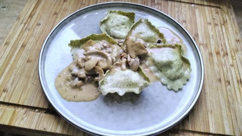 Grüne Ravioli liegen auf einem Teller, umgeben von einer cremigen Champignonsauce, auf einem Holzuntergrund.