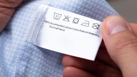Ein Waschetikett mit Waschsymbole zeigt "40°C", "Nicht bleichen", "Schonend trocknen", "Bügeln" und "Nicht chemisch reinigen". Es hängt an einem blauen Baumwollstoff.