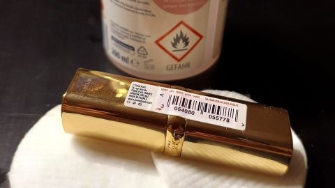 Nagellackentferner und Wattepads entfernen Klebeetiketten von einem goldenen Lippenstift. Text: "Gefahr", "Entferner ohne Aceton".