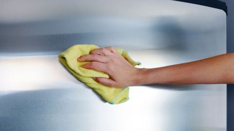 Eine Hand reinigt mit einem gelben Tuch sanft eine Edelstahloberfläche, vermutlich einen Kühlschrank, in einem modernen Raum.