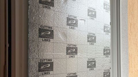 Alu-Trittschalldämmung bedeckt ein Fenster erkennbar an aufgedruckten „CUTTING LINES“ und reflektiert Licht im Innenbereich.