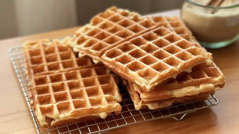 Belgische Waffeln liegen gestapelt auf einem Gitter, daneben steht ein Glas mit Teig auf einem Holztisch.