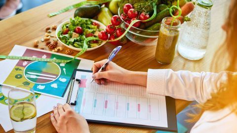 Eine Person schreibt an einem Tisch mit Ernährungsplänen, frischem Gemüse und Getränke; im Kontext von Ernährungswissenschaften.