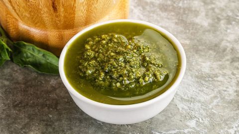 Basilikum-Pesto in einer weißen Schale ruht auf einem grauen Tisch neben einem hölzernen Mörser und Basilikumblättern.
