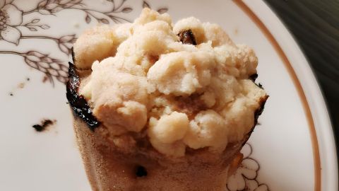 Muffin mit Streuseln steht auf floralem Porzellanteller, umgeben von Holzoberfläche. Keine sichtbare Schrift vorhanden.