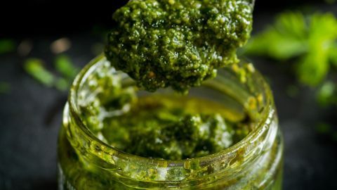 Ein Löffel hebt grünes Pesto aus einem Glas, umgeben von unscharfen grünen Kräutern im Hintergrund.