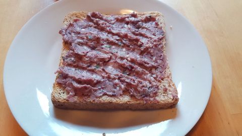 Ein Toast auf einem weißen Teller ist mit veganer Leberwurst bestrichen, auf einem hölzernen Tisch drapiert.